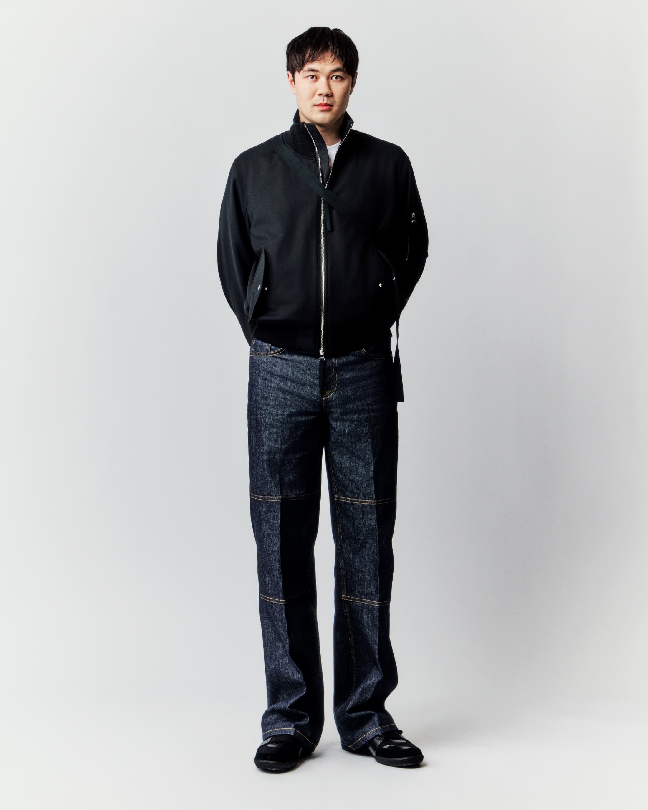 Full-length studio shot of an Asian man in a black jacket and dark denim jeans standing against a white background, fashion pose.