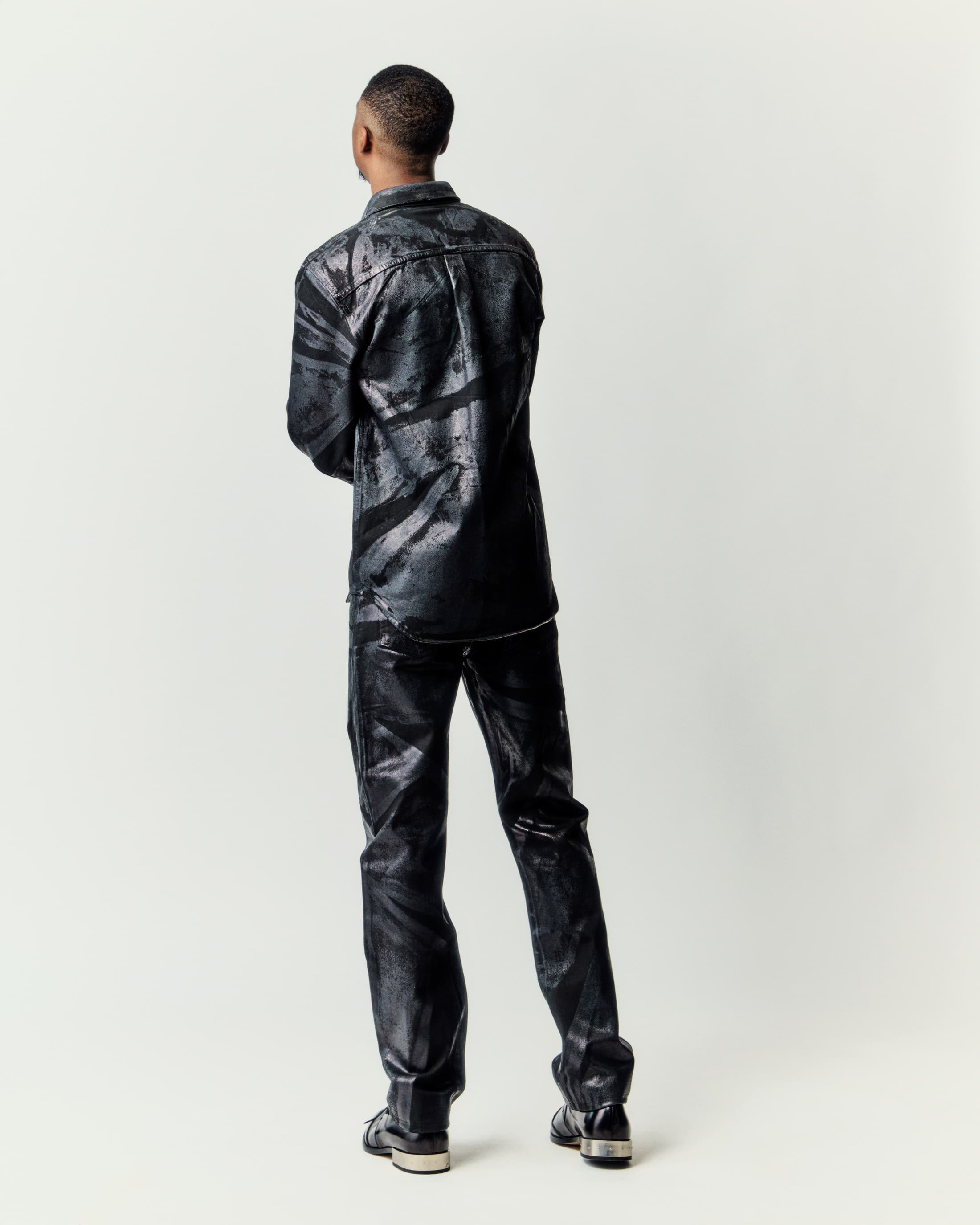 Back view of a black male in matching abstract-patterned shirt and pants standing in a bright studio with visible leather dress shoes.