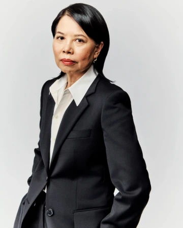 Portrait of a mature Asian woman in a dark suit, standing indoors against a bright neutral backdrop, radiating confidence and professionalism.