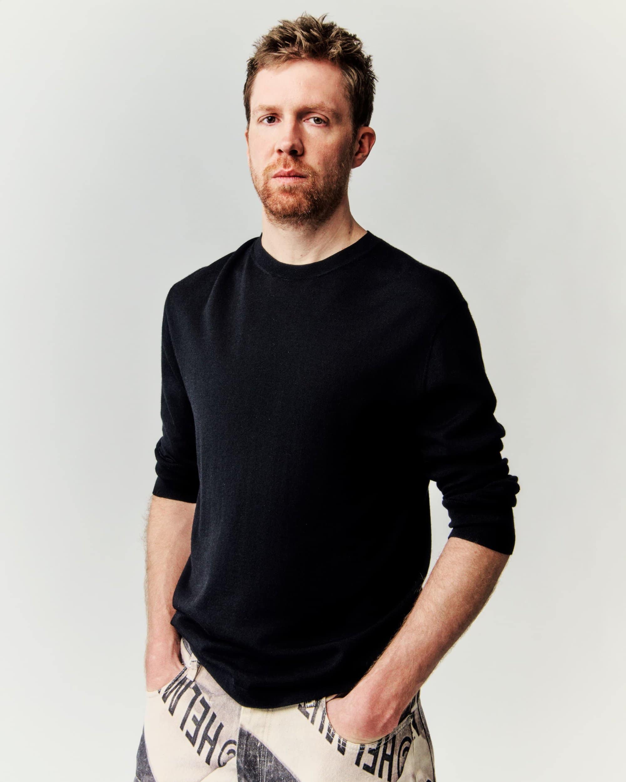 Close-up of an adult male with red hair and beard, wearing a black shirt and patterned pants, posing against a neutral backdrop.
