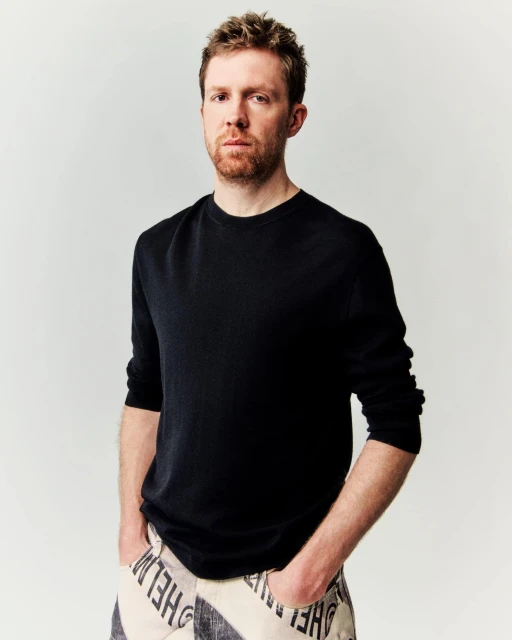 Close-up of an adult male with red hair and beard, wearing a black shirt and patterned pants, posing against a neutral backdrop.
