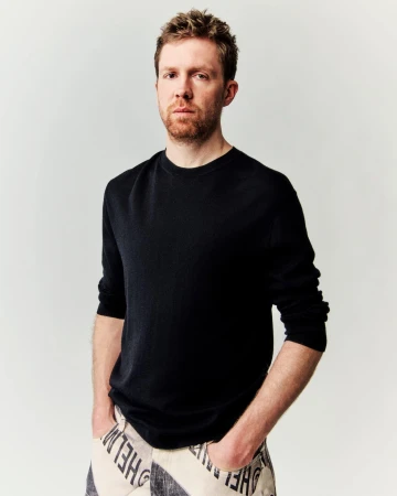 Close-up of an adult male with red hair and beard, wearing a black shirt and patterned pants, posing against a neutral backdrop.