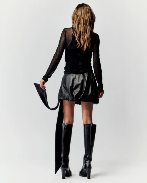 Back view of a female fashion model with long hair wearing a black top, leather skirt, and high-heeled boots against a white backdrop.