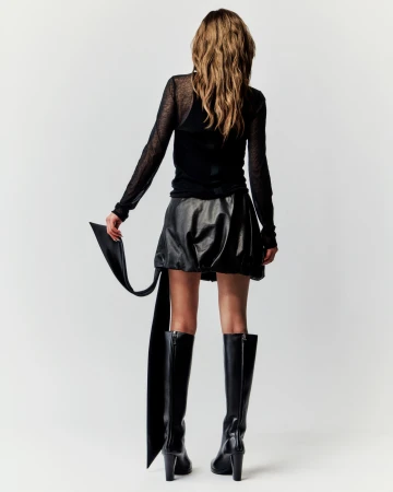 Back view of a female fashion model with long hair wearing a black top, leather skirt, and high-heeled boots against a white backdrop.
