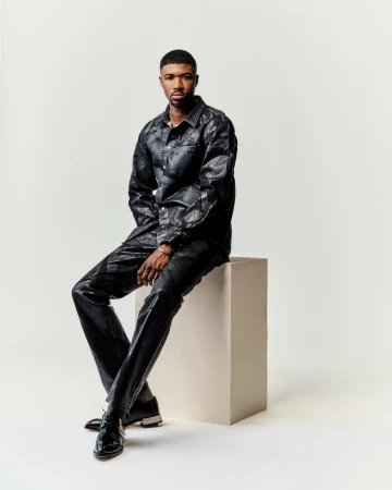 Man seated on a block wearing dark patterned shirt and trousers against a white seamless background