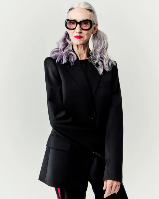 Elegant elderly woman with grey hair in pigtails wearing oversized black blazer and sunglasses against a light backdrop in studio setting.