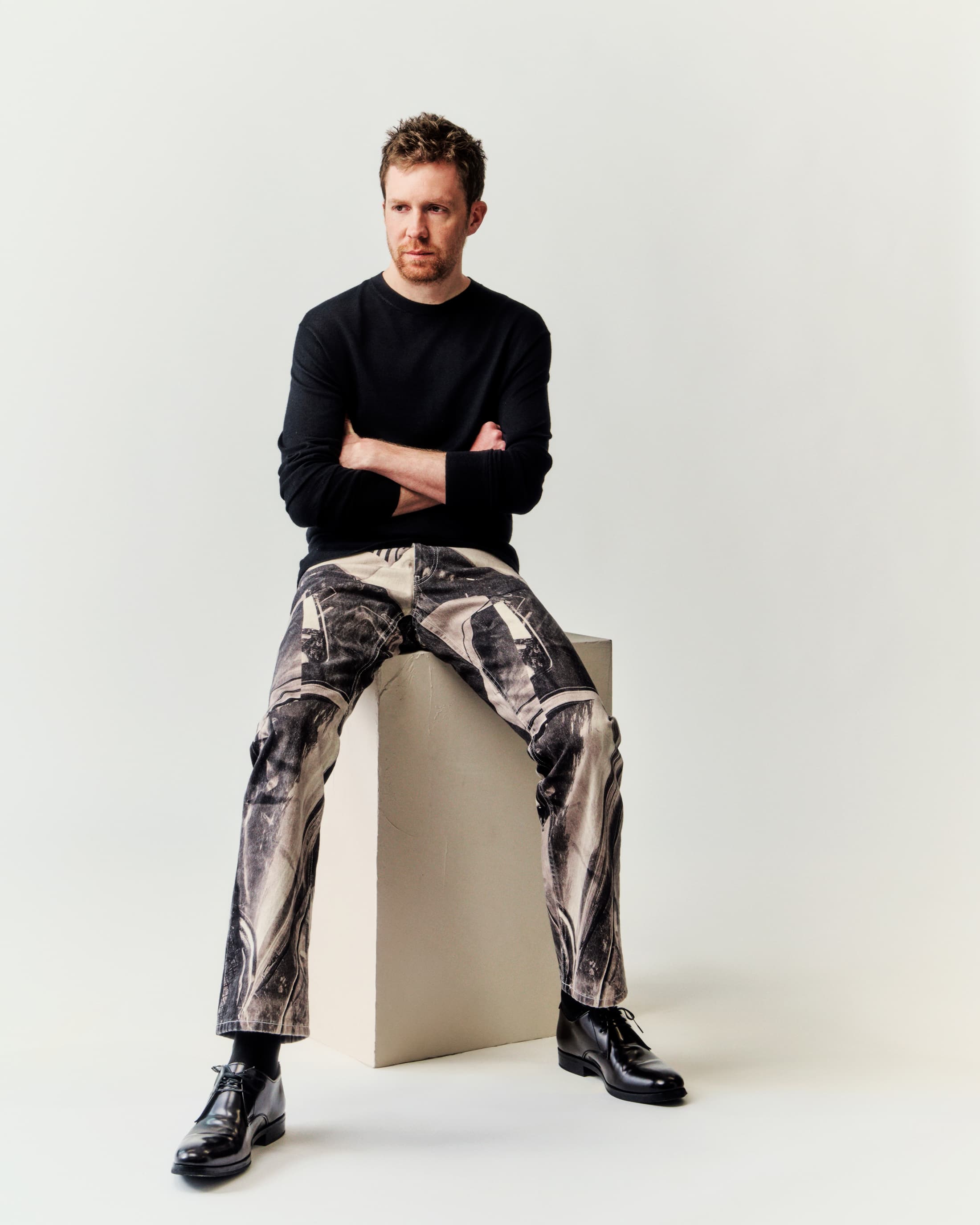 Portrait of a fair-skinned adult man with curly red hair. He is sitting indoors in a black top, patterned trousers, and black shoes.
