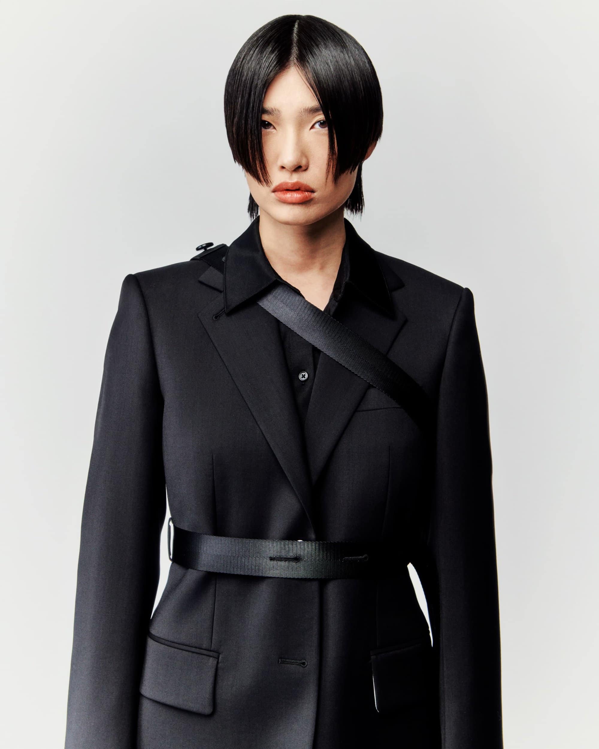 Portrait of a person with short black hair wearing a black blazer and shirt with a black belt, against a white background.