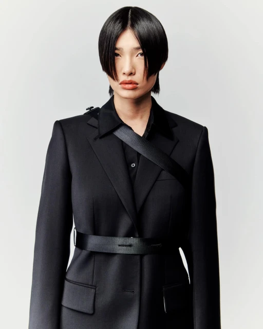 Portrait of a person with short black hair wearing a black blazer and shirt with a black belt, against a white background.
