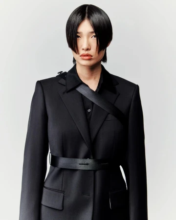Portrait of a person with short black hair wearing a black blazer and shirt with a black belt, against a white background.