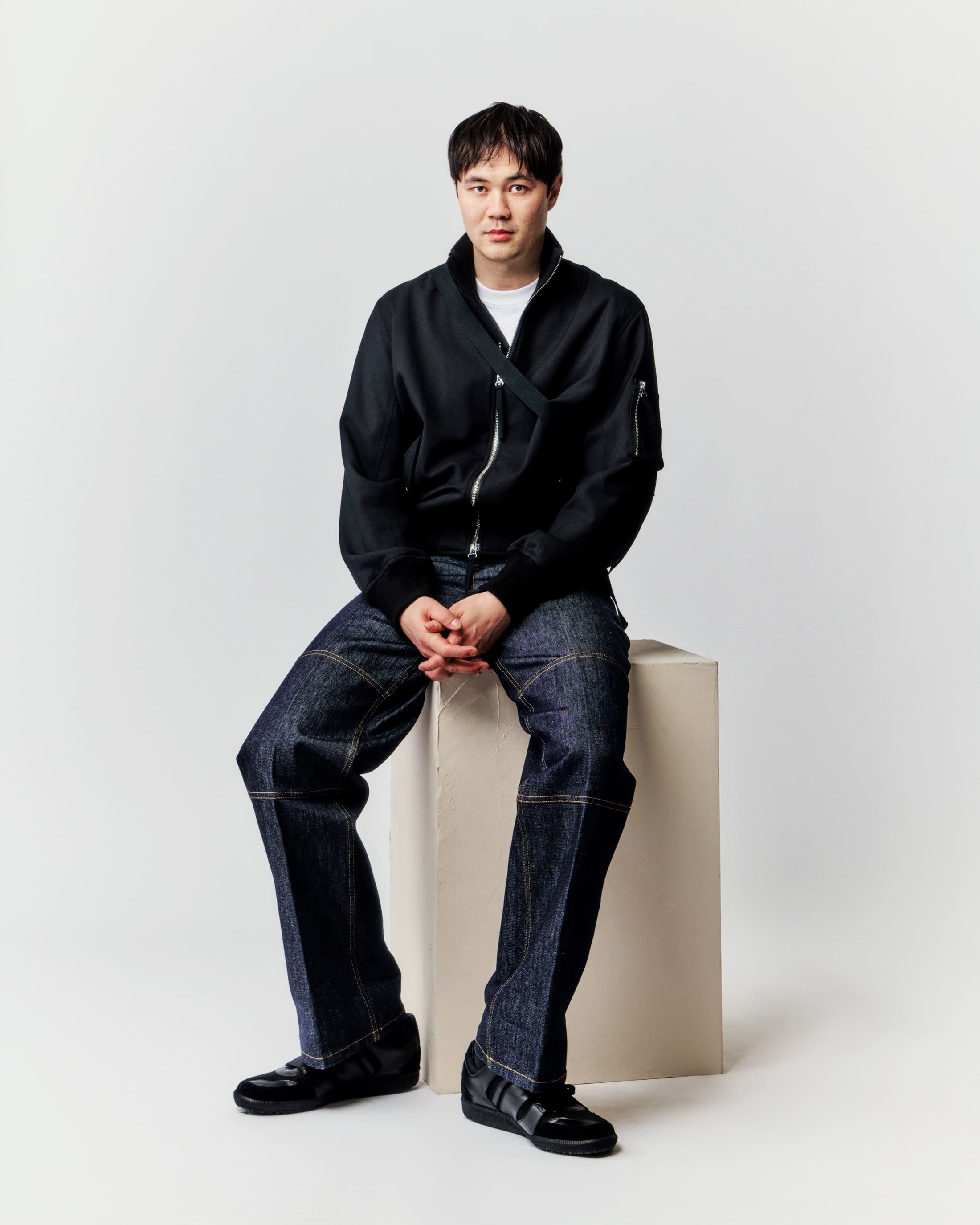 Full-length shot of a young adult male sitting on a white block, wearing a black bomber jacket, dark jeans, and black sneakers in a studio setting.