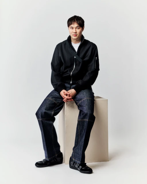 Full-length shot of a young adult male sitting on a white block, wearing a black bomber jacket, dark jeans, and black sneakers in a studio setting.