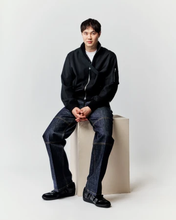Full-length shot of a young adult male sitting on a white block, wearing a black bomber jacket, dark jeans, and black sneakers in a studio setting.