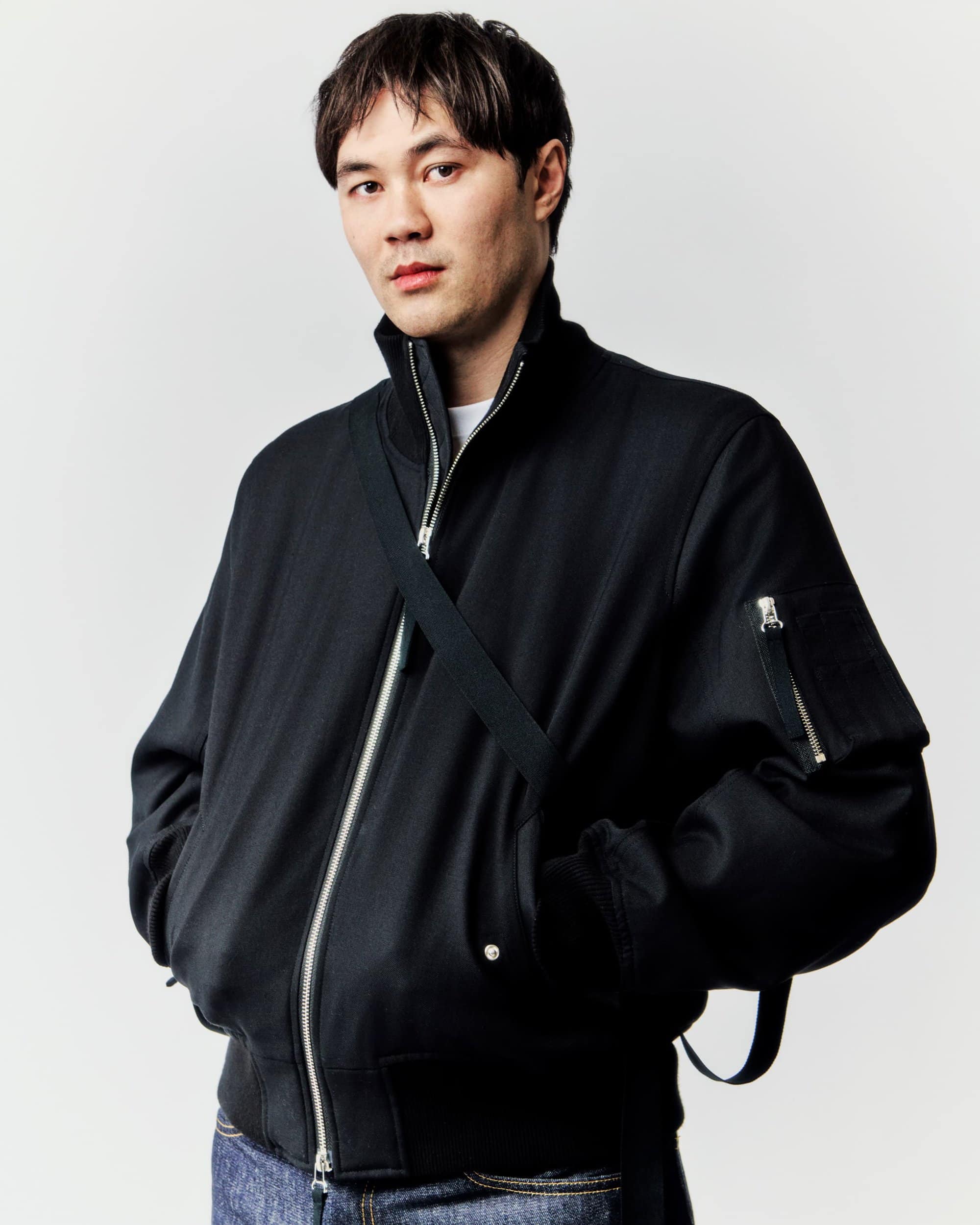 Studio portrait of an Asian man in a black bomber jacket and jeans with a cross-body bag strap, looking directly at the camera.