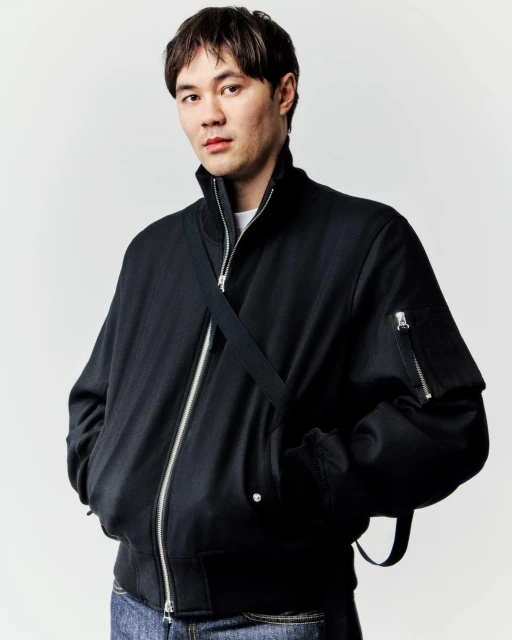 Studio portrait of an Asian man in a black bomber jacket and jeans with a cross-body bag strap, looking directly at the camera.