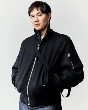 Studio portrait of an Asian man in a black bomber jacket and jeans with a cross-body bag strap, looking directly at the camera.