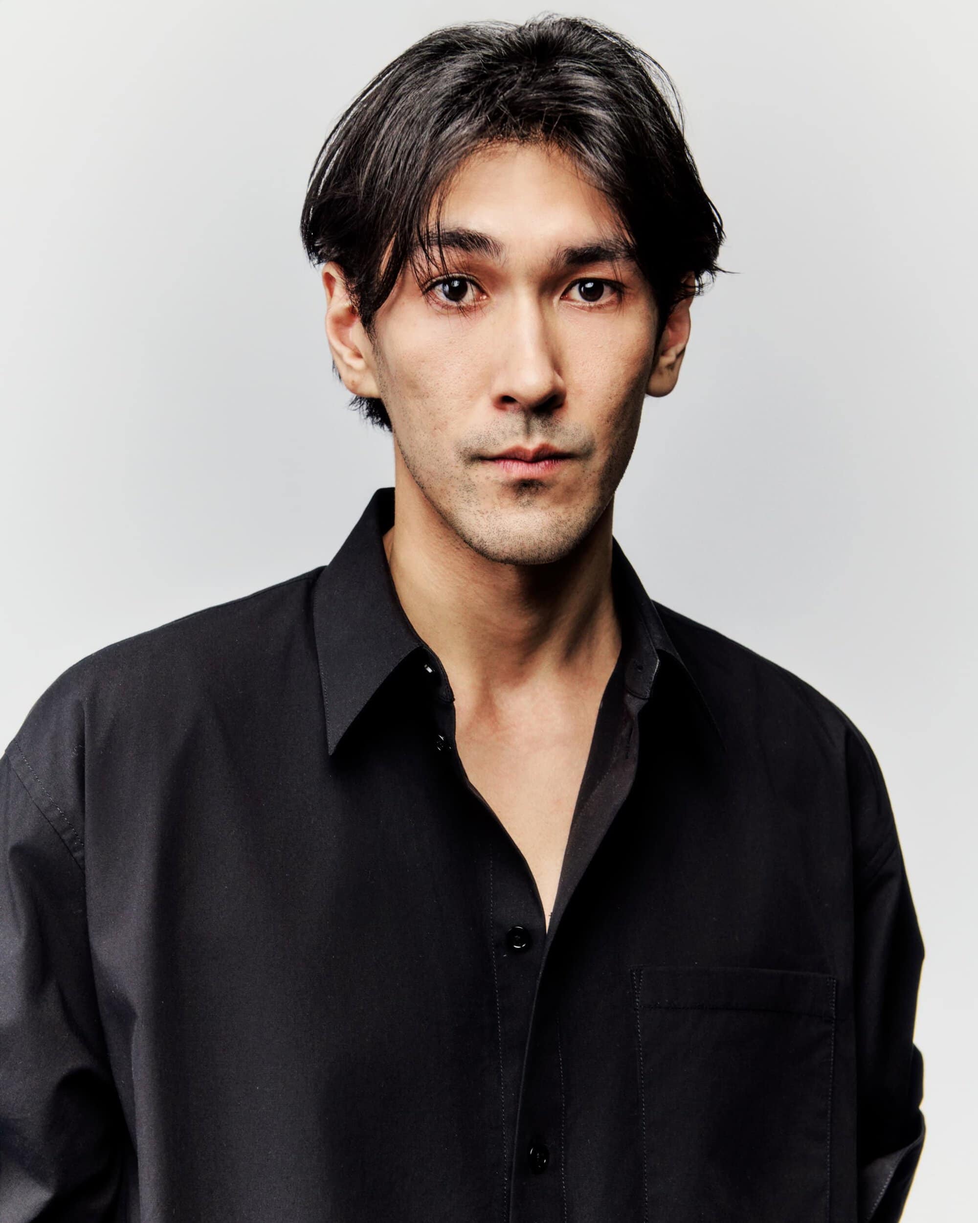Portrait of a young adult male with dark hair wearing a black button-down shirt against a neutral background in a photo studio.