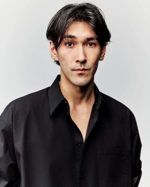 Portrait of a young adult male with dark hair wearing a black button-down shirt against a neutral background in a photo studio.