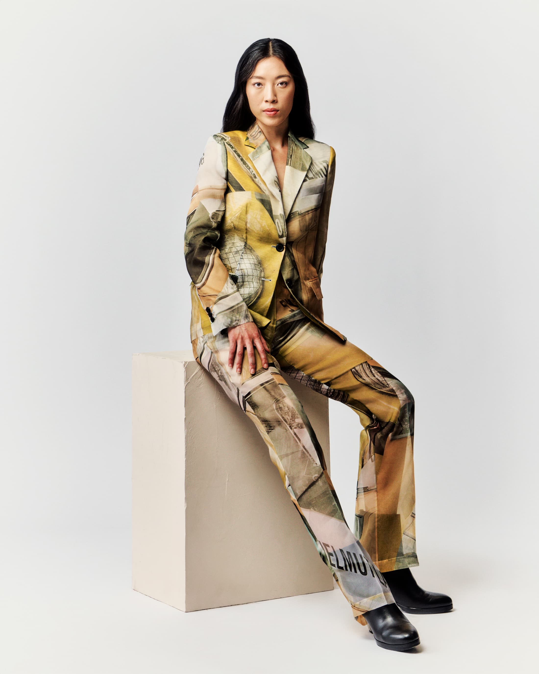 Woman wearing a patterned suit sits on a block in a photo studio, exuding an editorial fashion vibe with a neutral background.