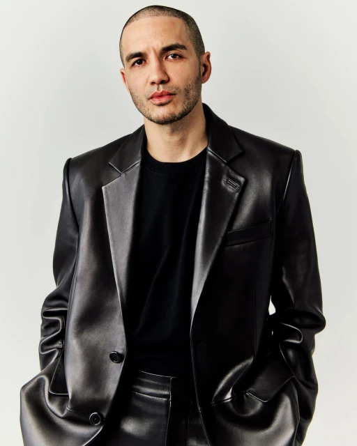 Close-up portrait of an adult male wearing a shiny black leather blazer and a black t-shirt against a neutral background in a studio shot.