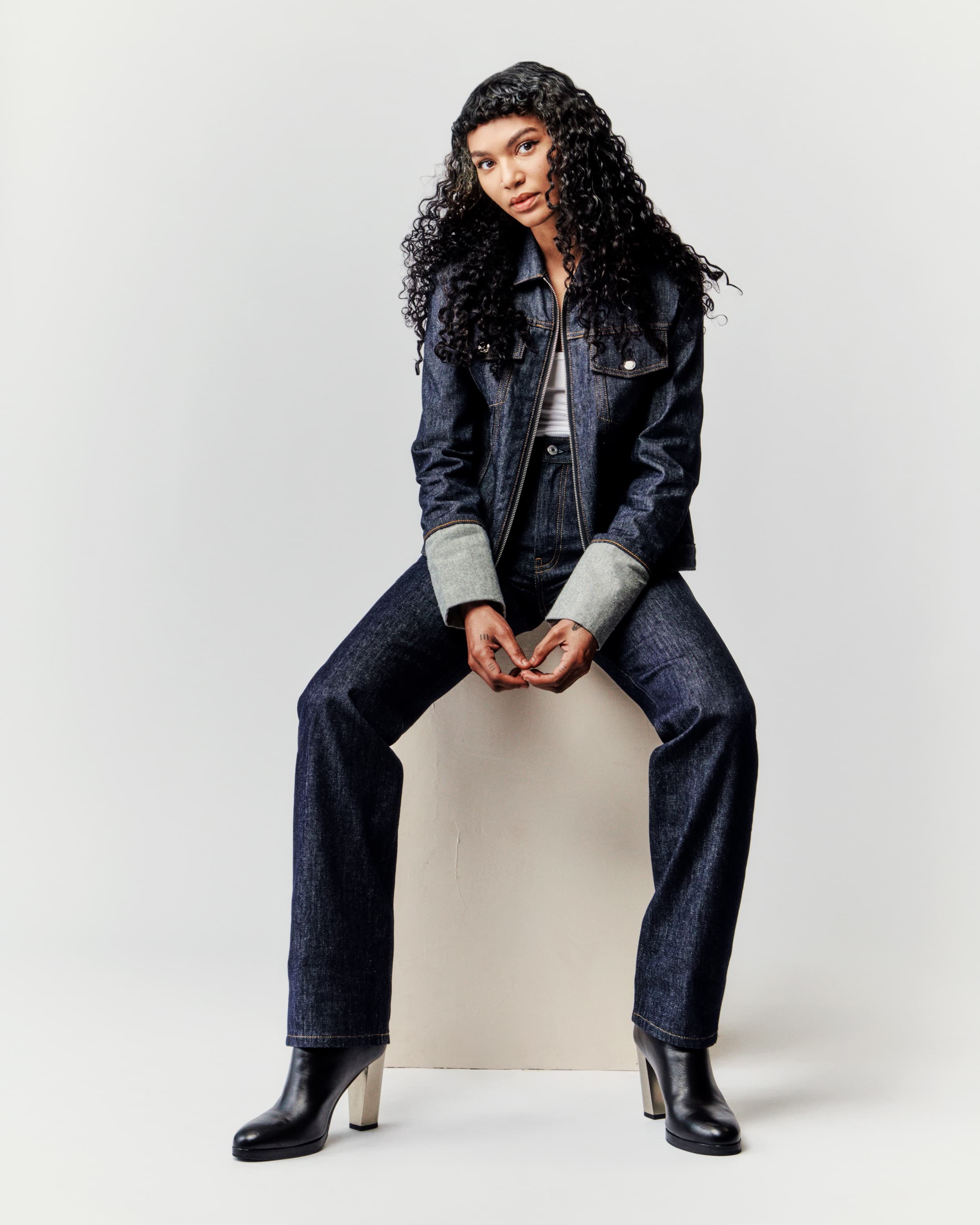 A young woman with curly hair sits on a white block wearing a denim jacket, matching wide-leg jeans, and black boots against a white background.