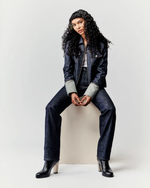 A young woman with curly hair sits on a white block wearing a denim jacket, matching wide-leg jeans, and black boots against a white background.