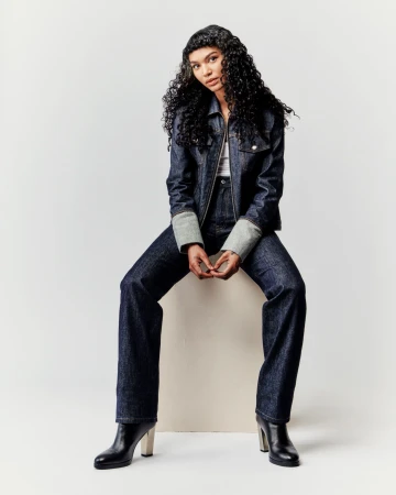 A young woman with curly hair sits on a white block wearing a denim jacket, matching wide-leg jeans, and black boots against a white background.