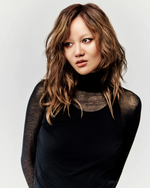 Portrait of an Asian woman with wavy brown and blonde hair wearing a black turtleneck top, set against a neutral grey background.