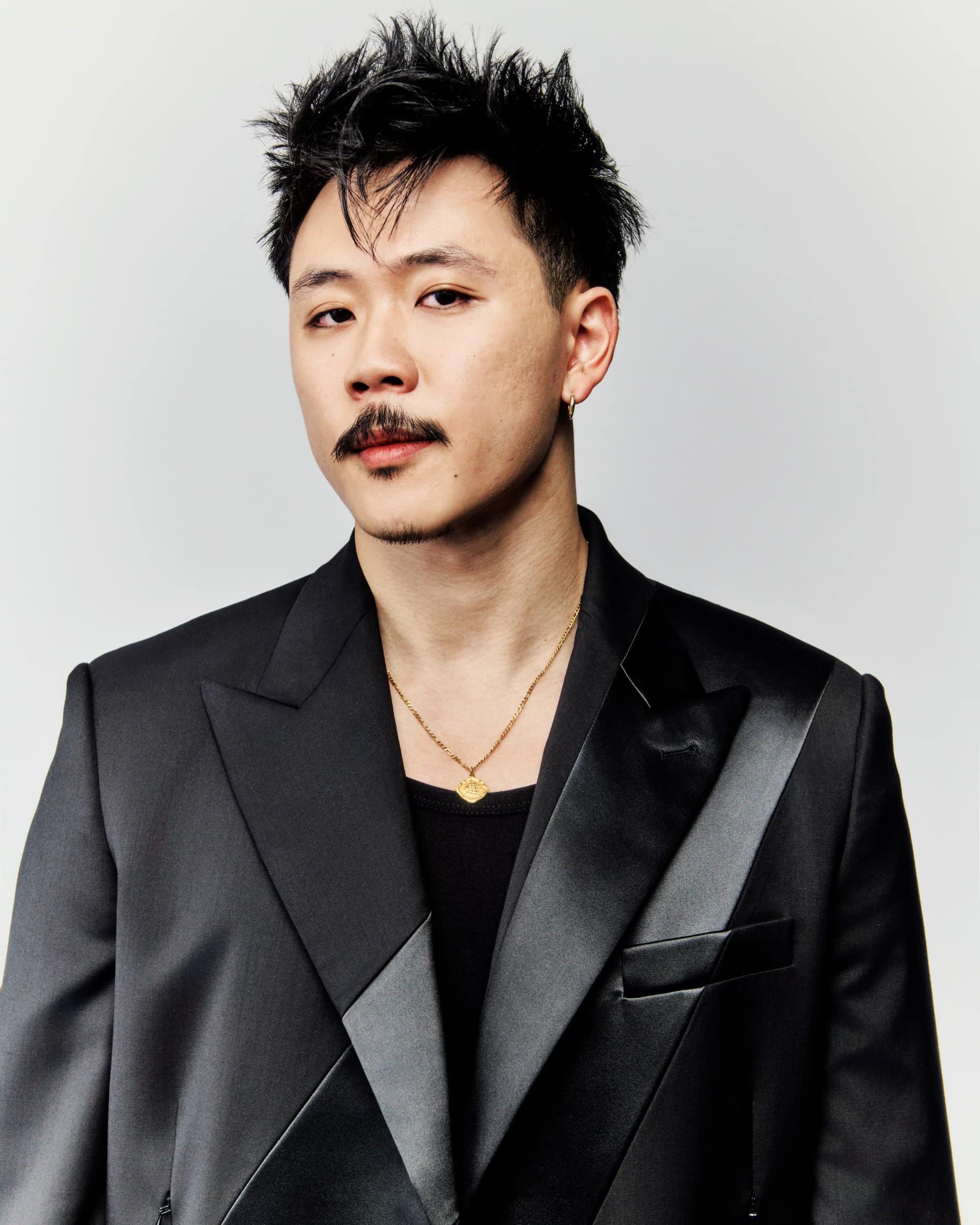 Portrait of an Asian man with a mustache wearing a black suit and gold necklace against a plain white background in a studio shot.