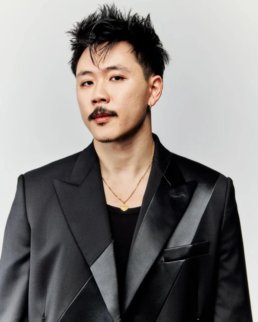 Portrait of an Asian man with a mustache wearing a black suit and gold necklace against a plain white background in a studio shot.