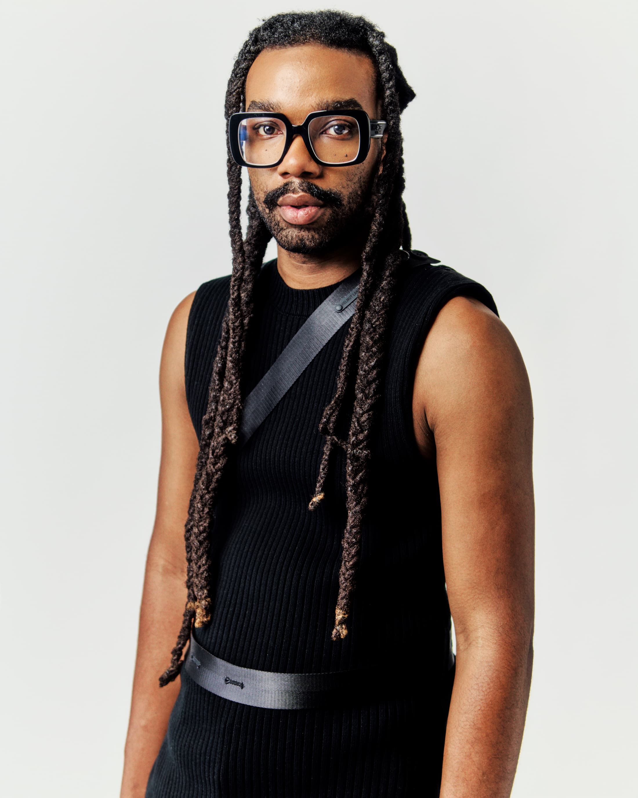 Headshot of a stylish Black adult male with braids, wearing glasses and a black sleeveless dress with shoulder and waist straps. 