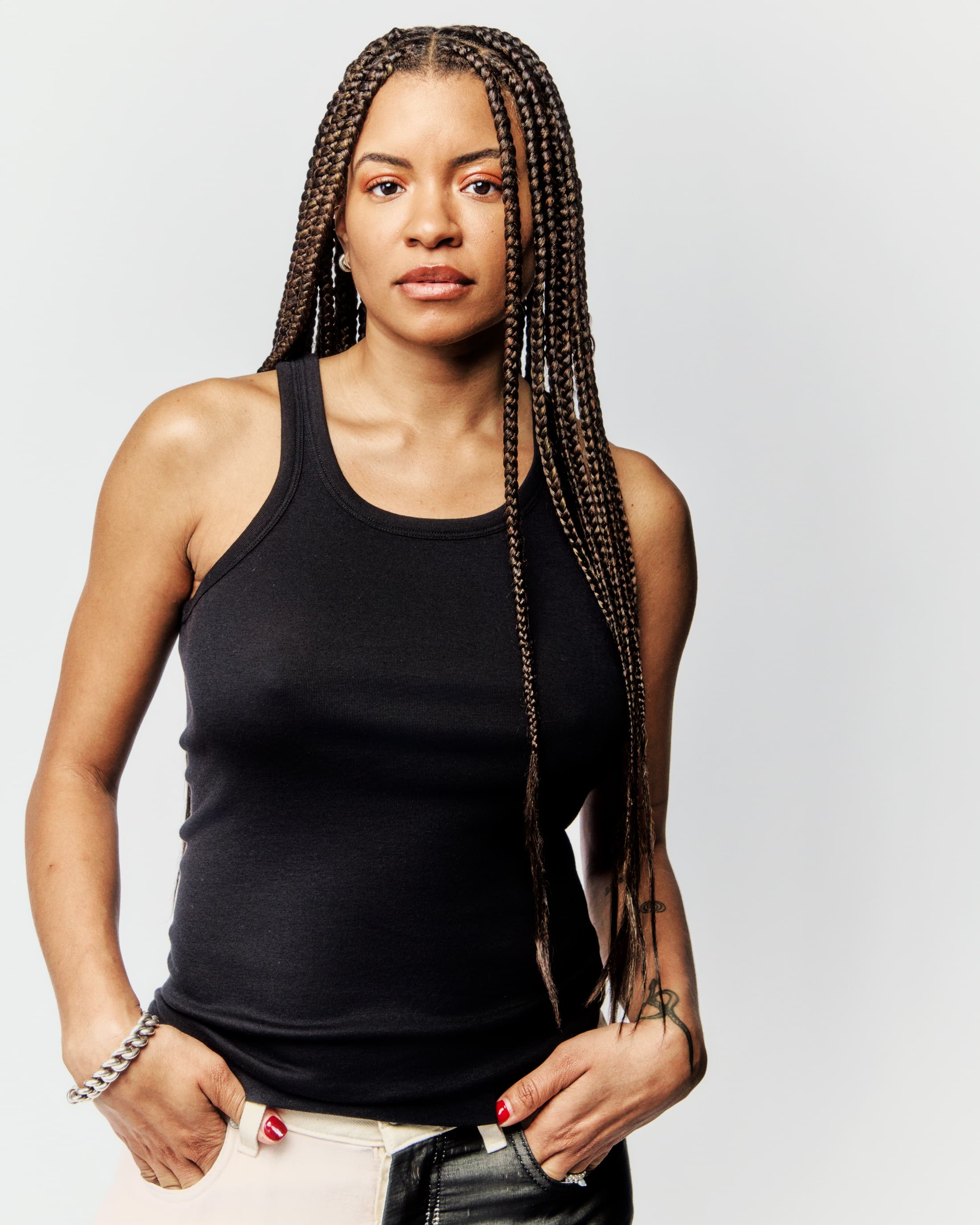 Portrait of a young adult woman with box braids, wearing a black tank top, and standing in a photo studio against a white background.