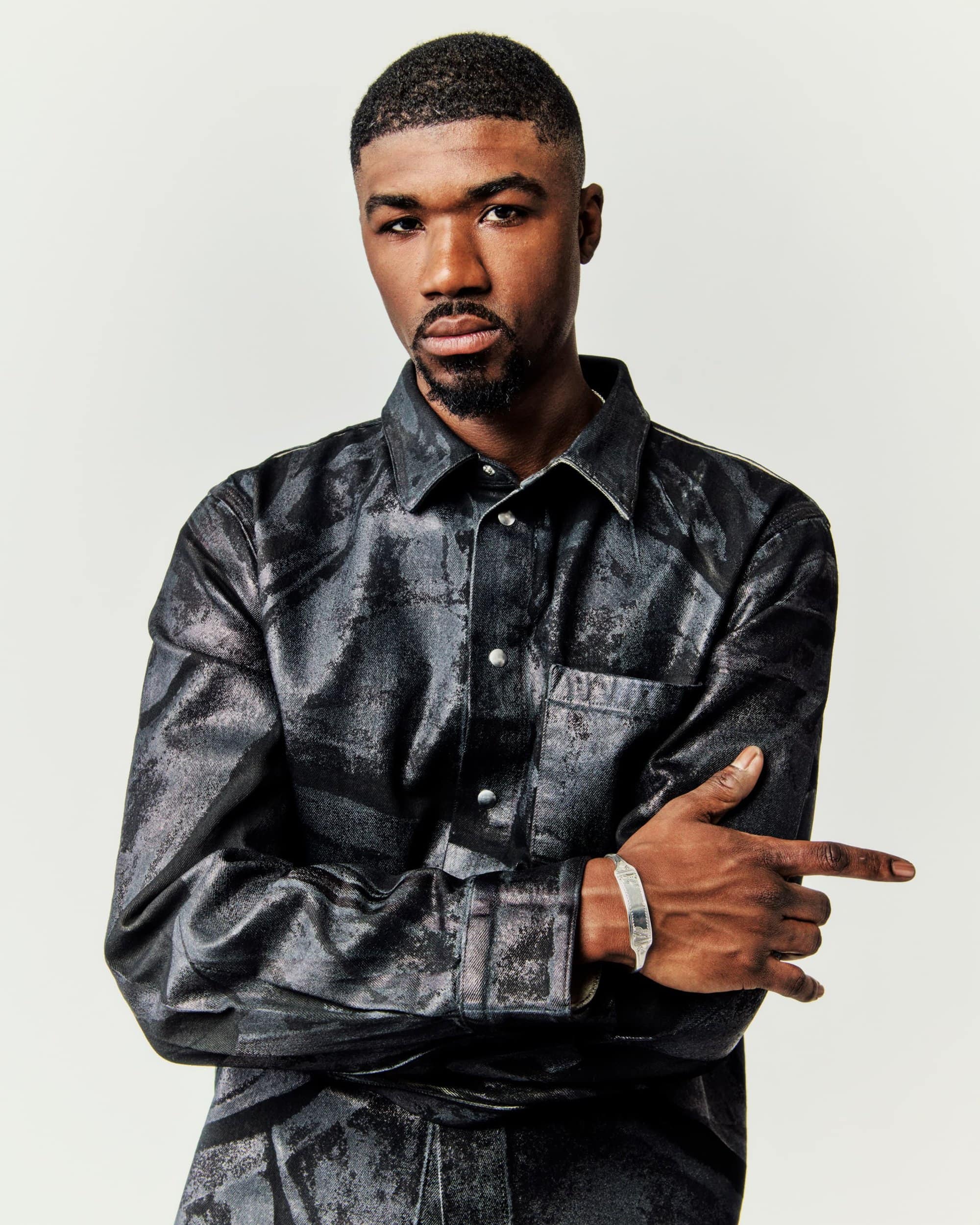 Portrait of a Black adult male with short hair and a goatee, wearing a patterned black shirt and silver bracelet in a studio setting.