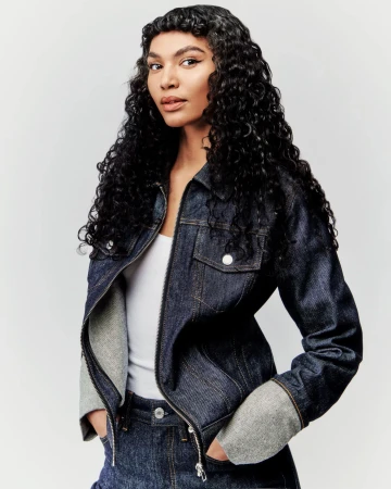 Model with long dark curly hair wearing a dark denim jacket and white top against a neutral light grey background.