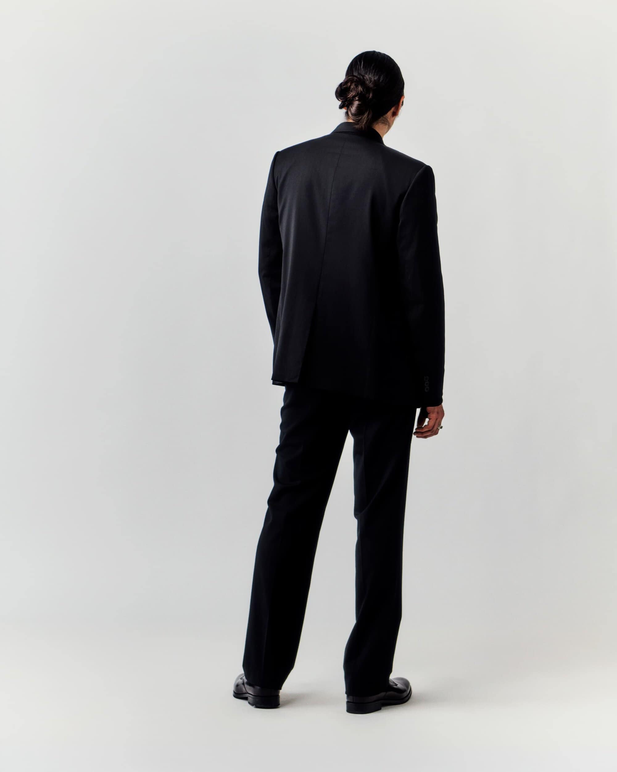 Back view of a slender adult male in a dark suit, featuring a single figure against a plain neutral backdrop in a studio setting.