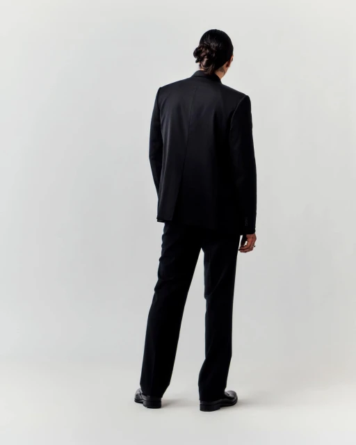 Back view of a slender adult male in a dark suit, featuring a single figure against a plain neutral backdrop in a studio setting.