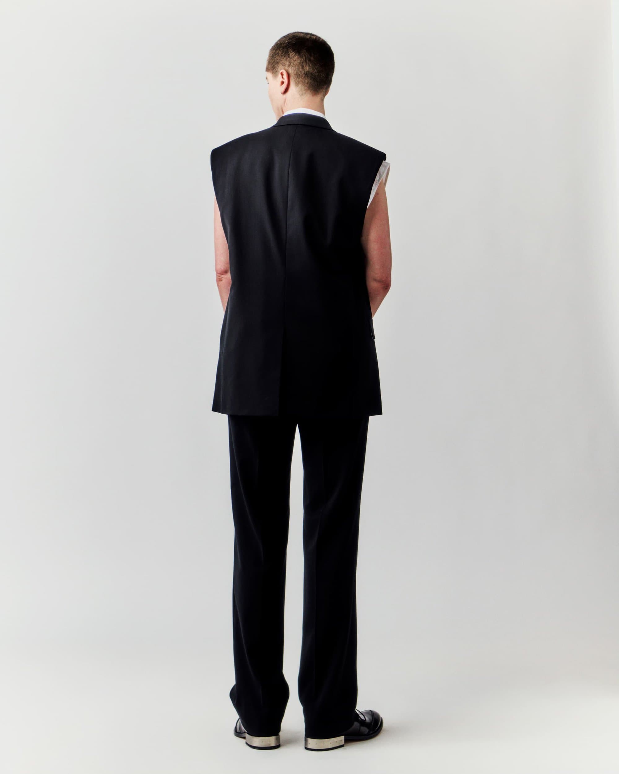Back view of an adult in a black vest and pants, standing in front of a plain white backdrop in a fashion or editorial shot.
