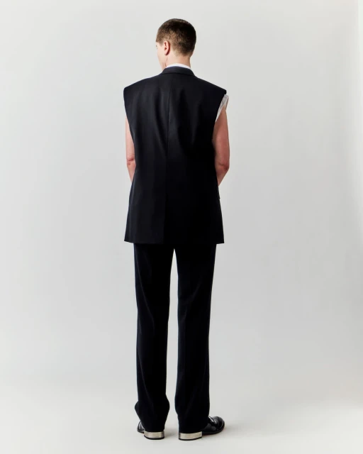 Back view of an adult in a black vest and pants, standing in front of a plain white backdrop in a fashion or editorial shot.