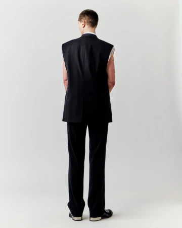Back view of an adult in a black vest and pants, standing in front of a plain white backdrop in a fashion or editorial shot.