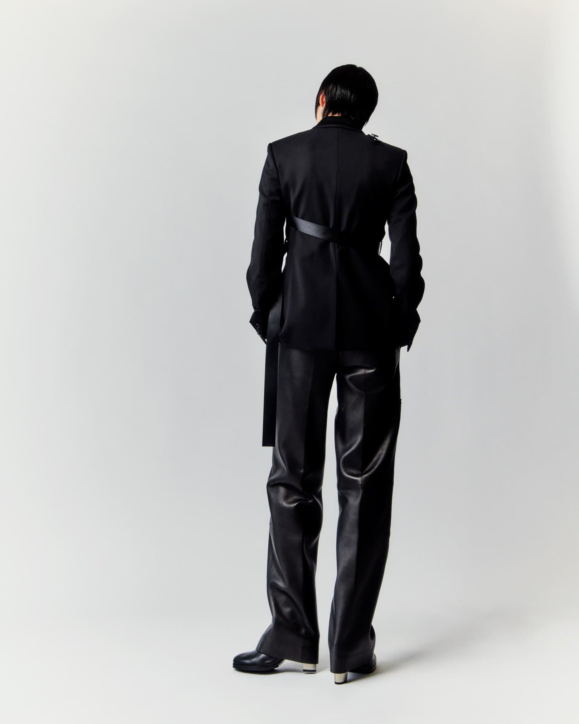 Person in black suit and leather pants poses against white backdrop, facing away, showcasing avant-garde fashion and style.