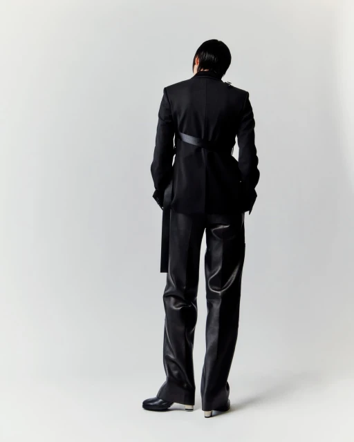 Person in black suit and leather pants poses against white backdrop, facing away, showcasing avant-garde fashion and style.