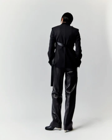 Person in black suit and leather pants poses against white backdrop, facing away, showcasing avant-garde fashion and style.