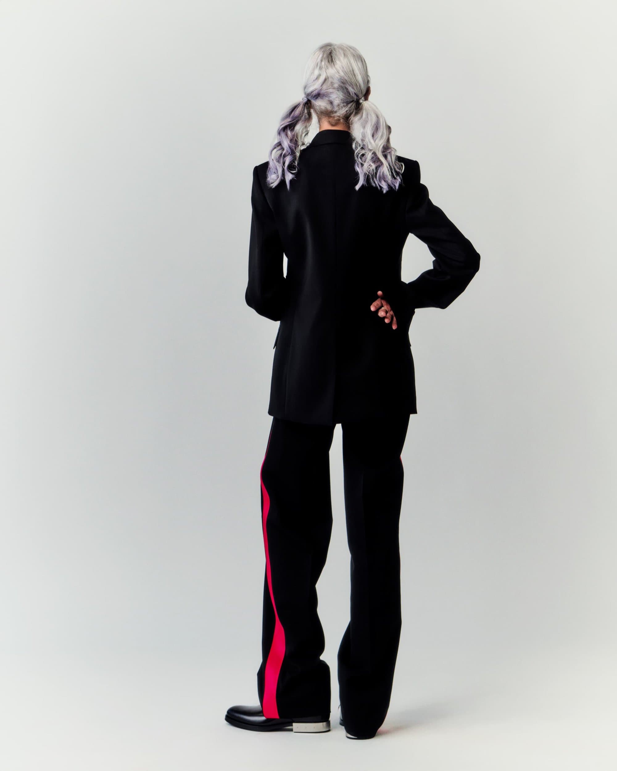 Fashionable adult female posing with back turned, wearing a black blazer, black pants with red stripes, and black shoes in a studio.