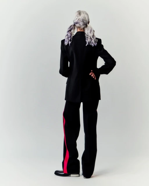 Fashionable adult female posing with back turned, wearing a black blazer, black pants with red stripes, and black shoes in a studio.