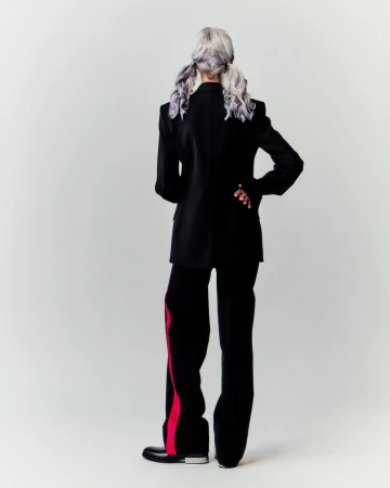 Fashionable adult female posing with back turned, wearing a black blazer, black pants with red stripes, and black shoes in a studio.
