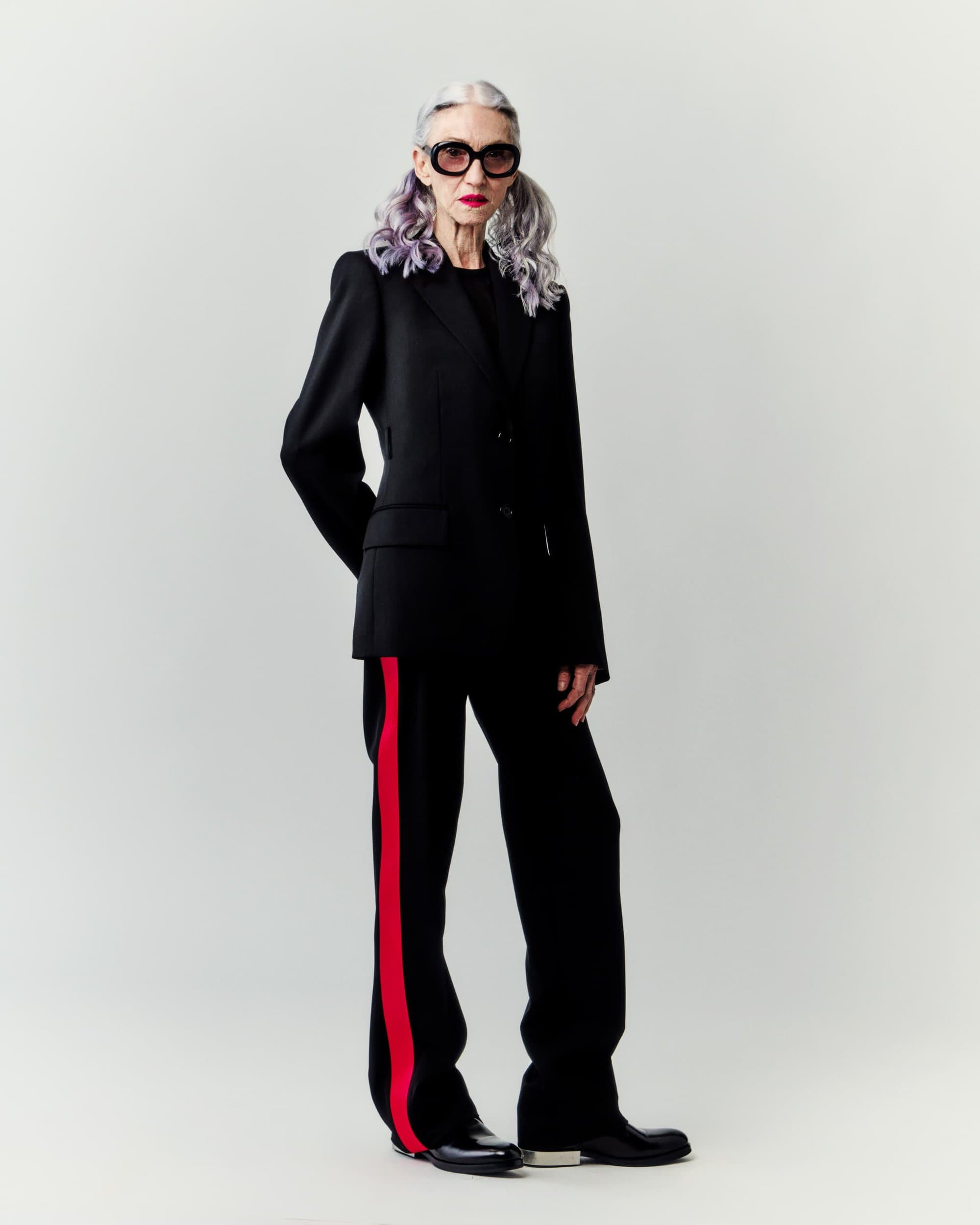 Mature woman in a black pantsuit with a red stripe and dark glasses, poses in a studio with soft lighting against a bright backdrop.