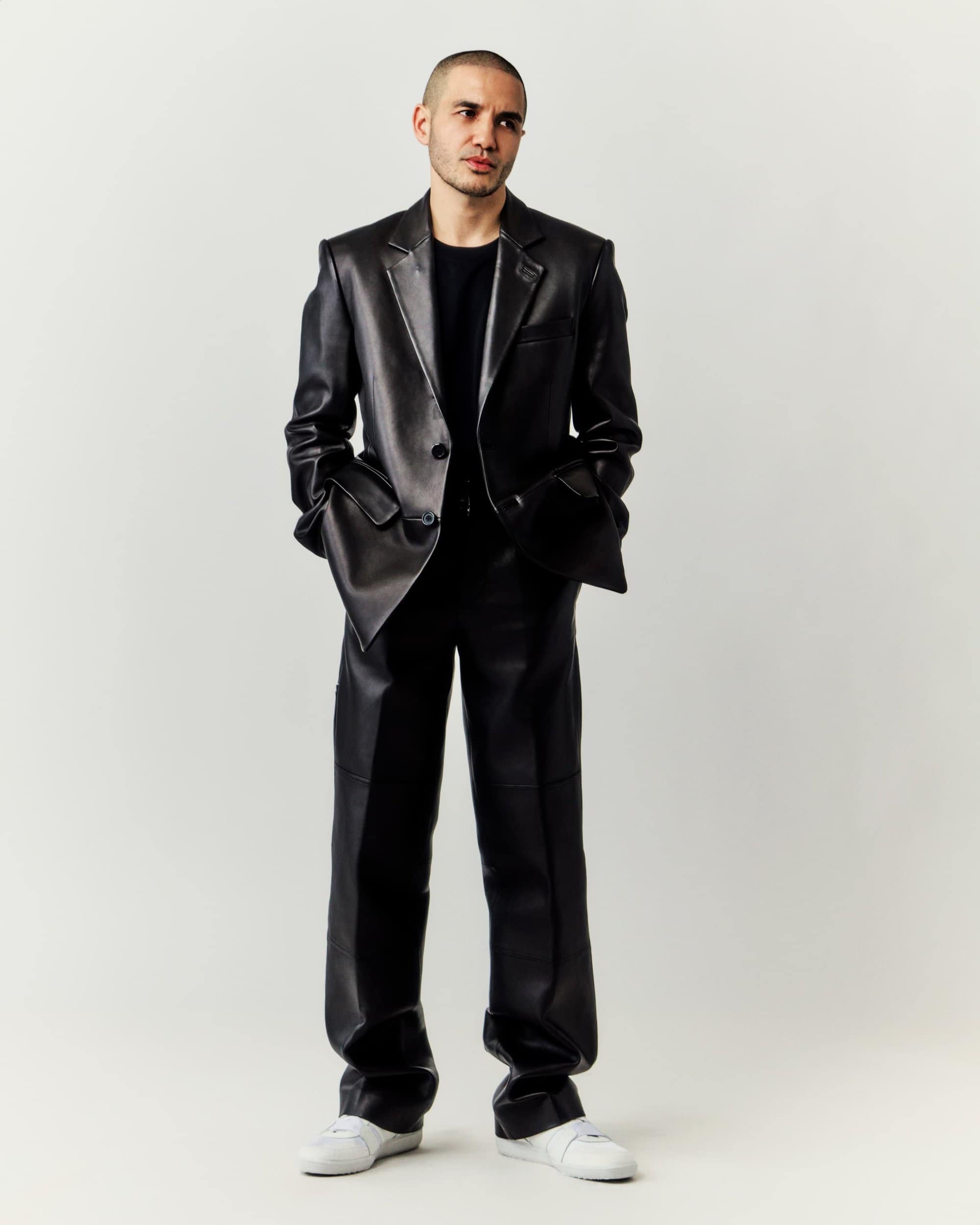 Full-length portrait of a young adult male in a black leather suit and white sneakers, standing against a neutral background in studio lighting.