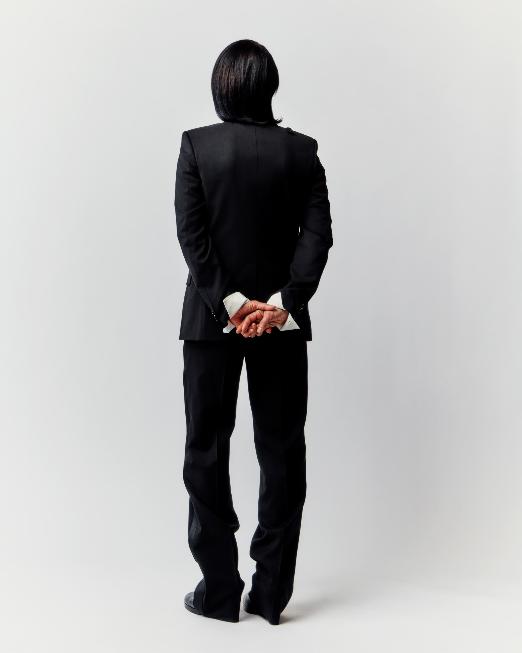 Adult male in formal black suit stands with hands clasped behind his back against a stark white backdrop in a studio shot.