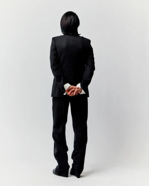 Adult male in formal black suit stands with hands clasped behind his back against a stark white backdrop in a studio shot.