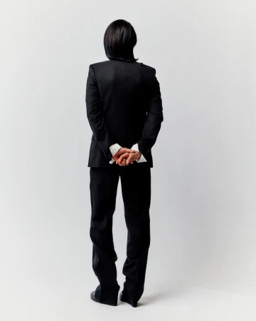 Model from behind wearing a black suit jacket and trousers with hands clasped against a light grey background.